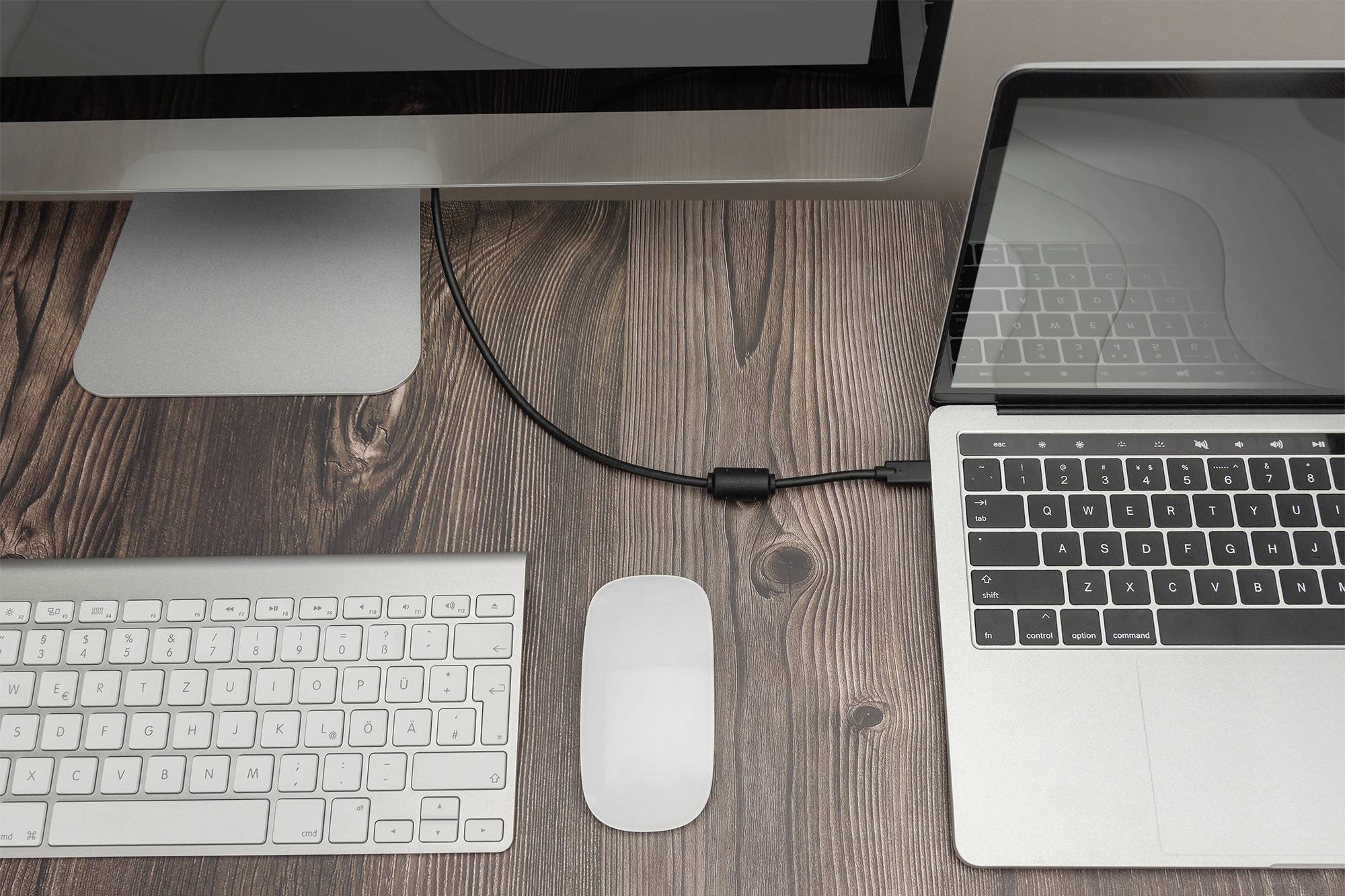 Ein Laptop und ein Desktop-Computer sind auf einem Holztisch verbunden. Eine Tastatur und Maus liegen daneben.