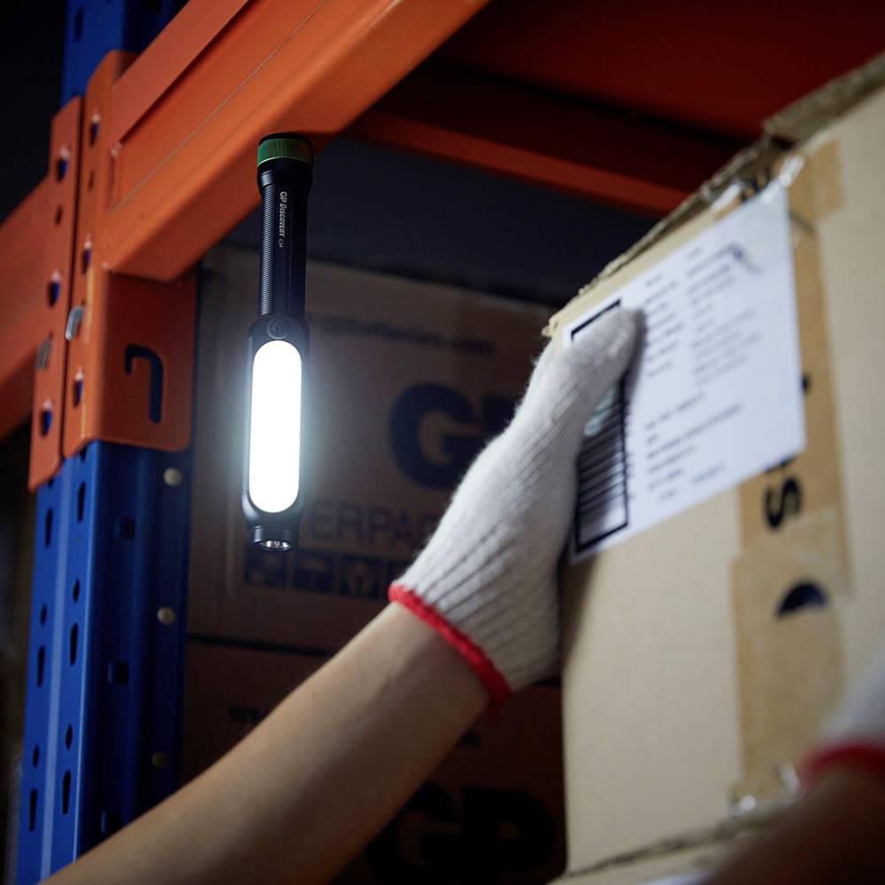 Eine Hand mit einem weißen Handschuh hält einen Karton in einem Lager. Eine Taschenlampe hängt an einem Regal und beleuchtet die Szene.