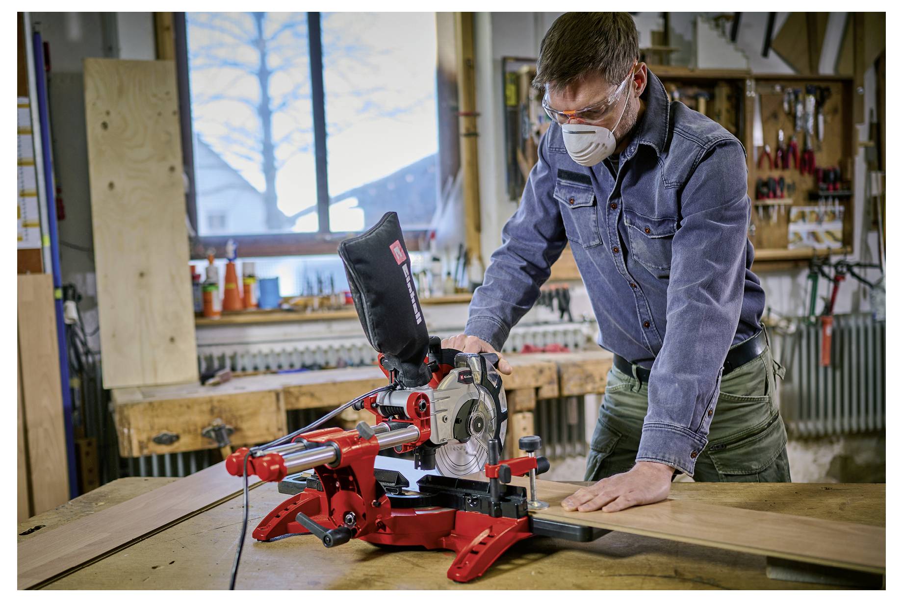 Ein Mann mit einer Maske bedient eine rote Gehrungssäge in einer Werkstatt und schneidet ein Holzbrett auf einer Werkbank.