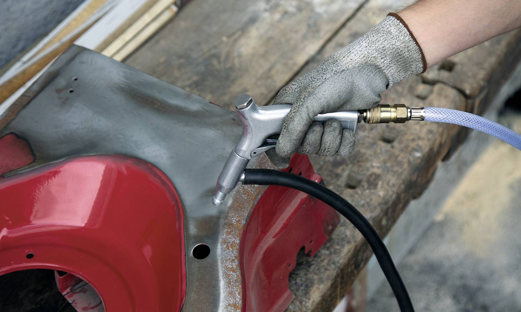 Eine Person mit grauen Arbeitshandschuhen sandstrahlt ein Metallstück auf einer Holzoberfläche; der Lack ist teilweise entfernt.