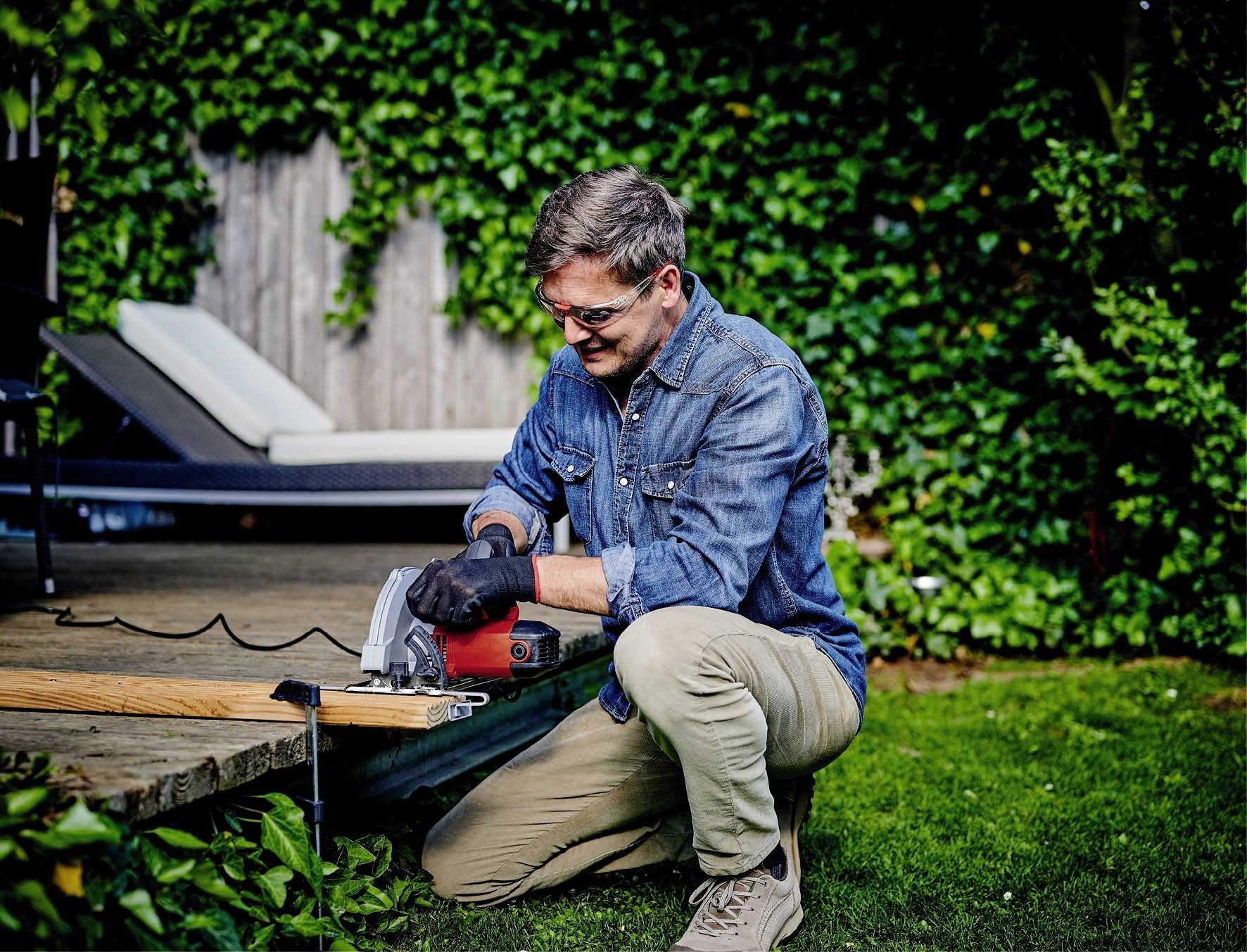 Ein Mann in Arbeitskleidung kniet in einem Garten und schneidet ein Holzbrett mit einer elektrischen Säge. Im Hintergrund sind Pflanzen und eine Liege.