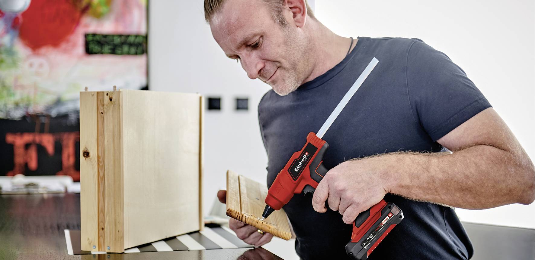 Ein Mann befestigt ein Holzbrett mit einem Akku-Bohrschrauber an einer Box. Im Hintergrund ist unscharf ein buntes Bild sichtbar.