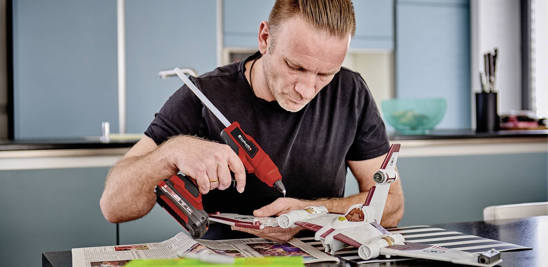 Ein Mann arbeitet konzentriert mit einem Akkuschrauber an einem Modellflugzeug auf einem Tisch in einer modernen Küche.