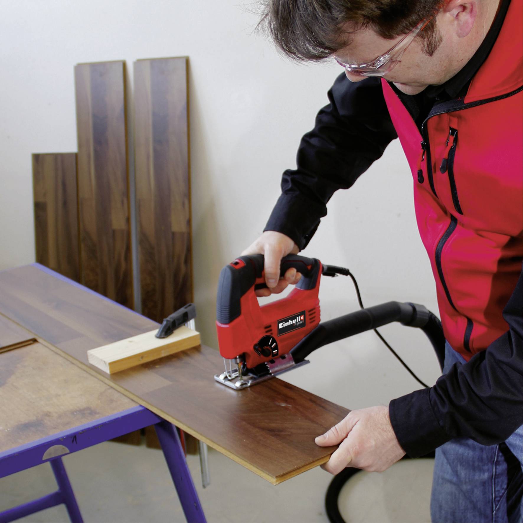 Eine Person in roter Arbeitsjacke schneidet ein Holzbrett mit einer roten Elektrosäge auf einem Arbeitstisch.