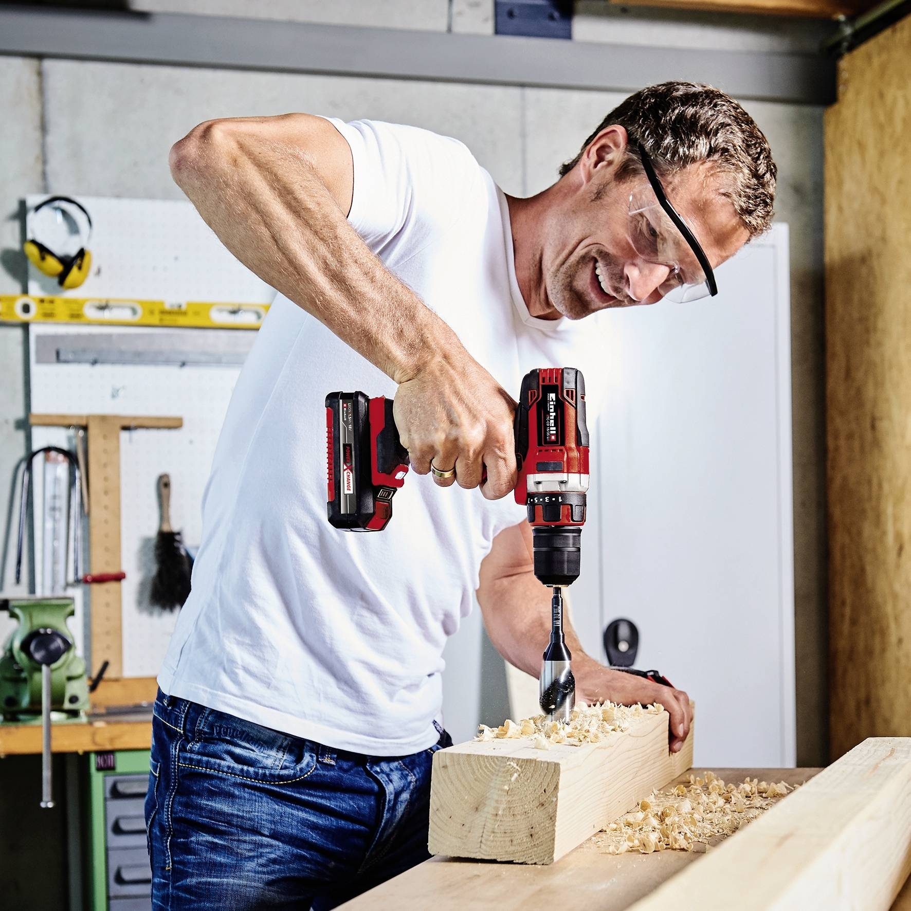 Ein Mann in Schutzbrille und weißem T-Shirt bohrt mit einem Akkubohrer ein Loch in ein Holzbrett in einer Werkstattumgebung.