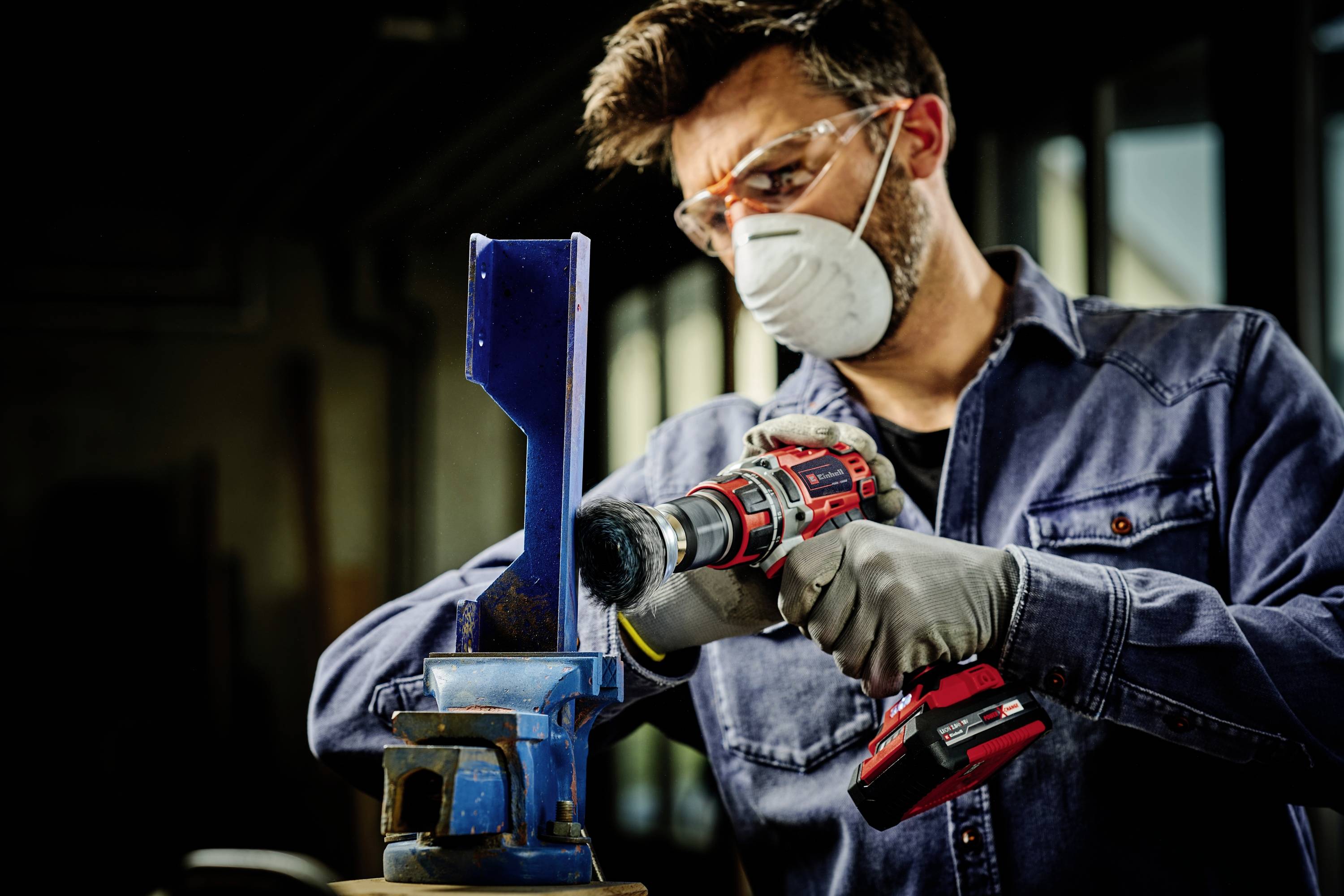 Ein Mann mit Schutzbrille und Maske schleift ein Metallstück mit einem Bohrschleifer in einer Werkstatt.