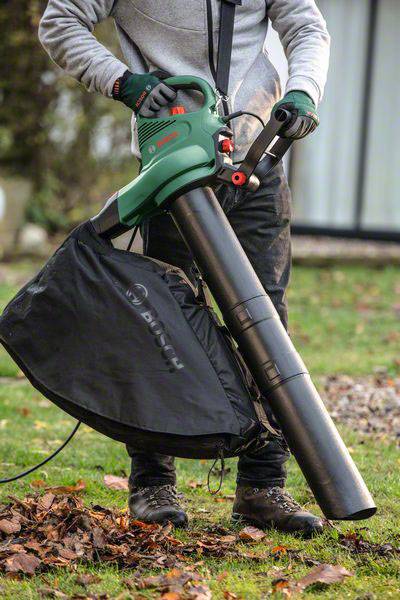 Eine Person benutzt einen Laubbläser, um Blätter im Garten aufzusaugen. Der Laubbläser ist grün und mit einem schwarzen Sammelbehälter ausgestattet.