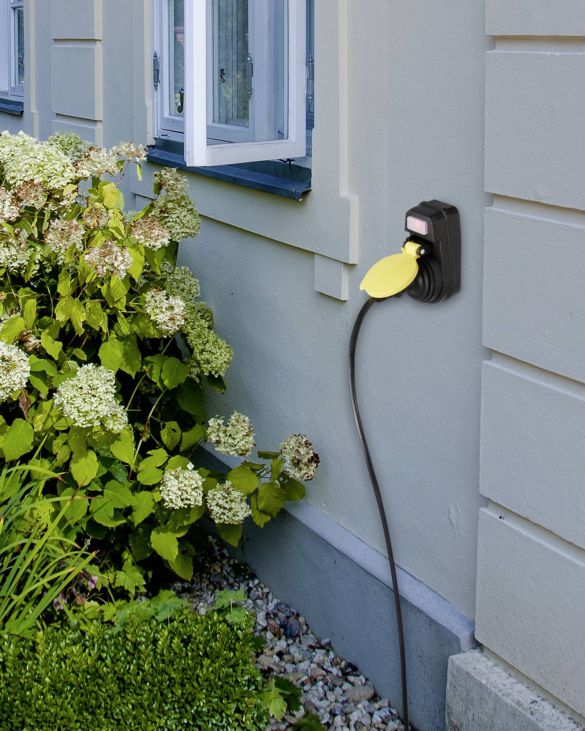 Außensteckdose an einer Hauswand mit einem eingesteckten Kabel im Garten, umgeben von Hortensien und Kieselsteinen.