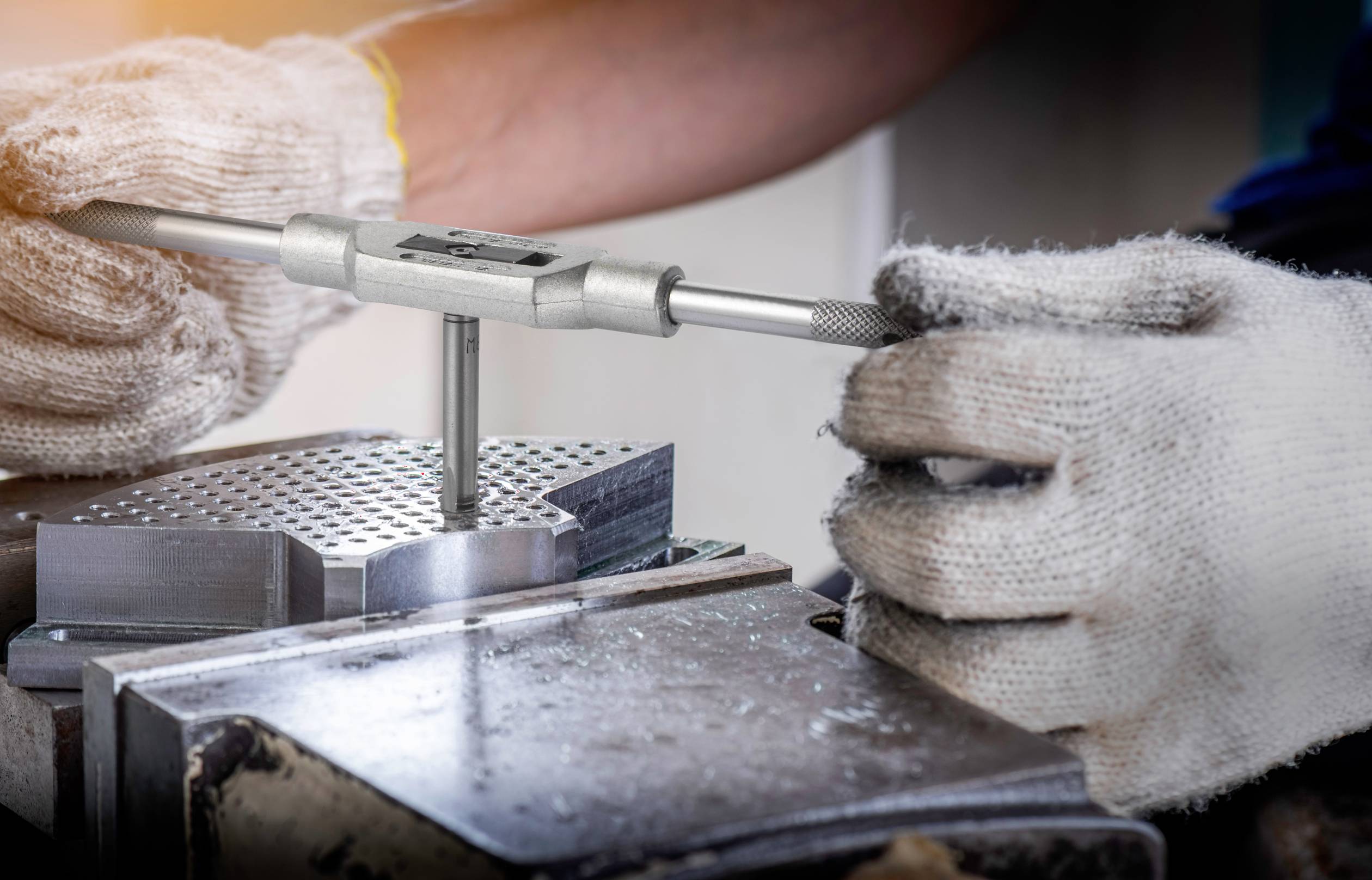 Arbeiter trägt Sicherheitshandschuhe und justiert ein Gewindeschneidewerkzeug in einer Werkstatt. Metallische Werkstücke auf Werkbank.