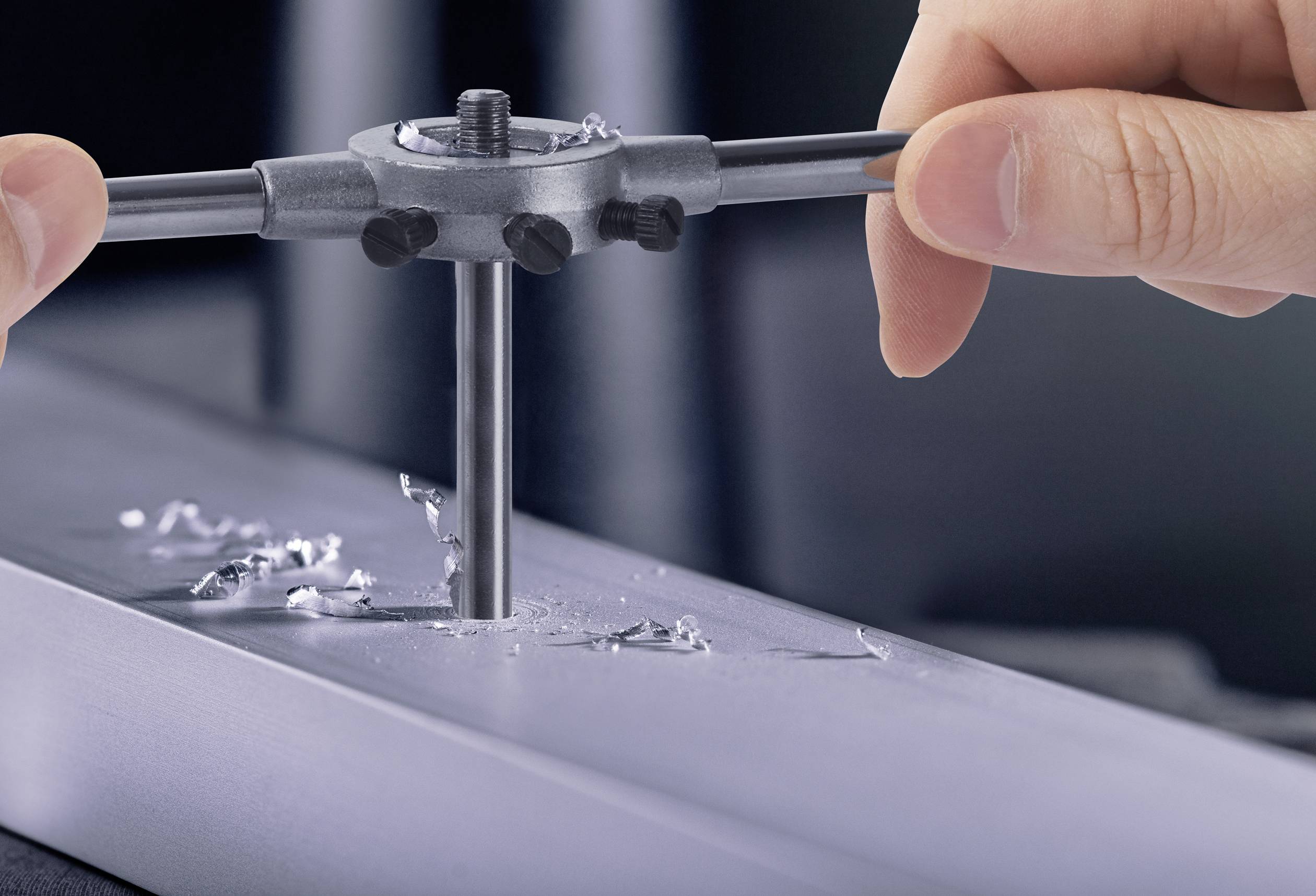 Zwei Hände nutzen ein Gewindeschneider-Werkzeug, um präzise Gewinde in ein Metallstück zu schneiden. Metallspäne sind sichtbar.