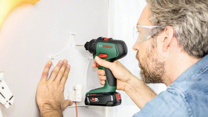 Ein Mann bohrt mit einem kabellosen Bohrer in die Wand, trägt Schutzbrille. Er hält eine Halterung gegen die Wand.