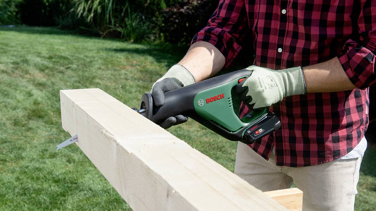 Eine Person in kariertem Hemd und Handschuhen sägt mit einer elektrischen Säge ein Holzbrett im Garten.