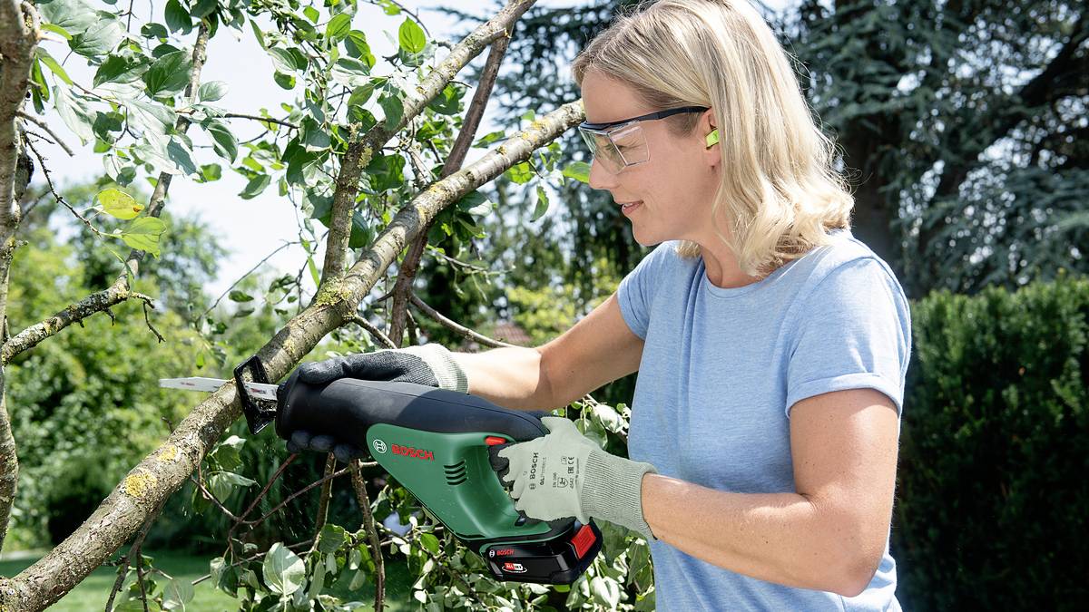 Eine Frau mit Gehörschutz und Schutzbrille verwendet eine Akku-Säge, um einen Ast im Garten zu kürzen.