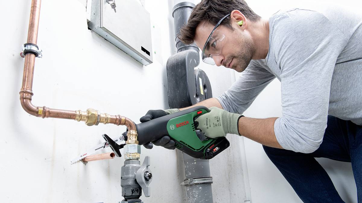 Ein Mann mit Schutzbrille und Ohrstöpseln verwendet eine elektrische Säge, um ein Kupferrohr in einem industriellen Umfeld zu schneiden.