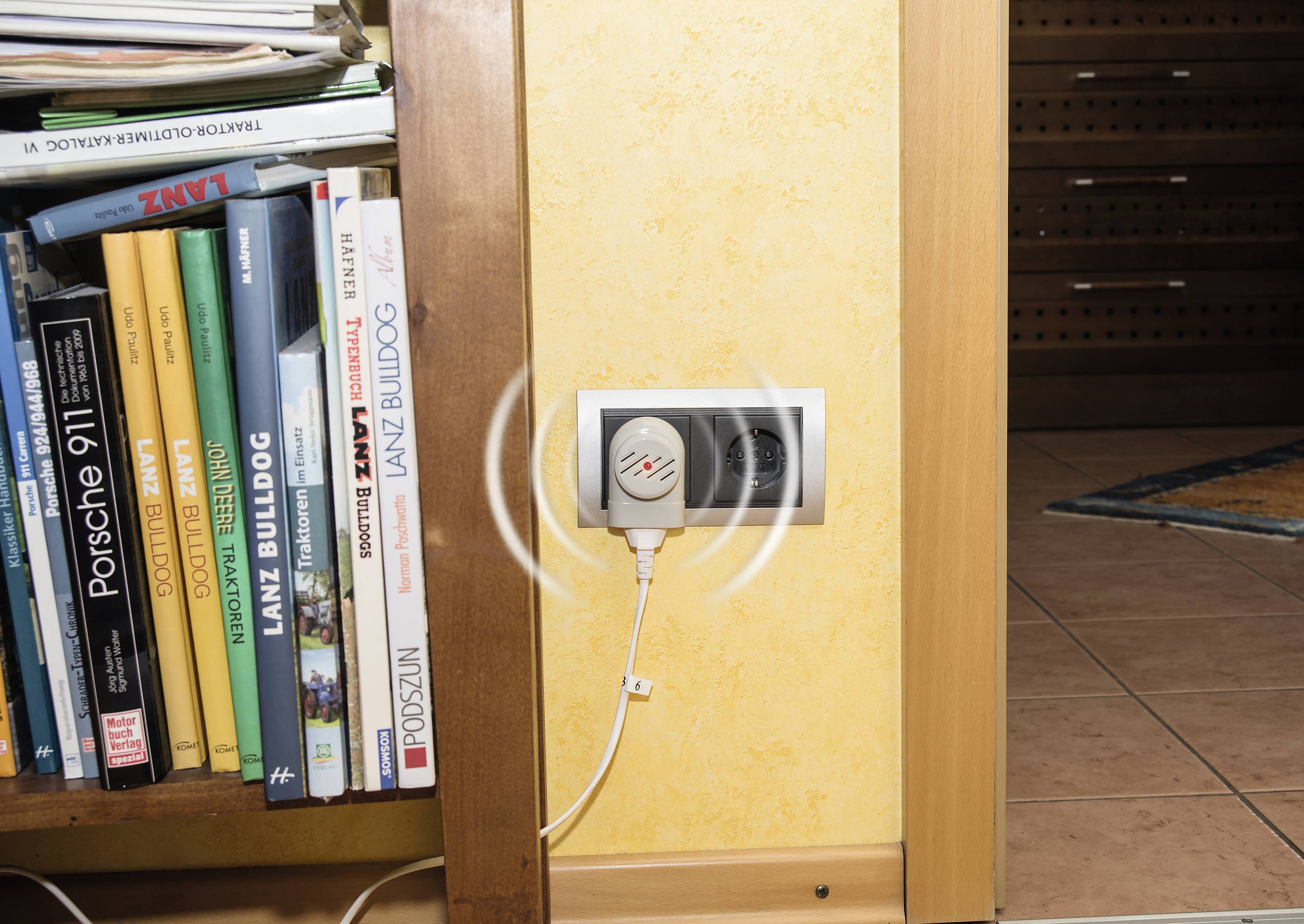 Bücherregal voller Bücher neben einer Wandsteckdose, in die ein elektrisches Gerät eingesteckt ist.