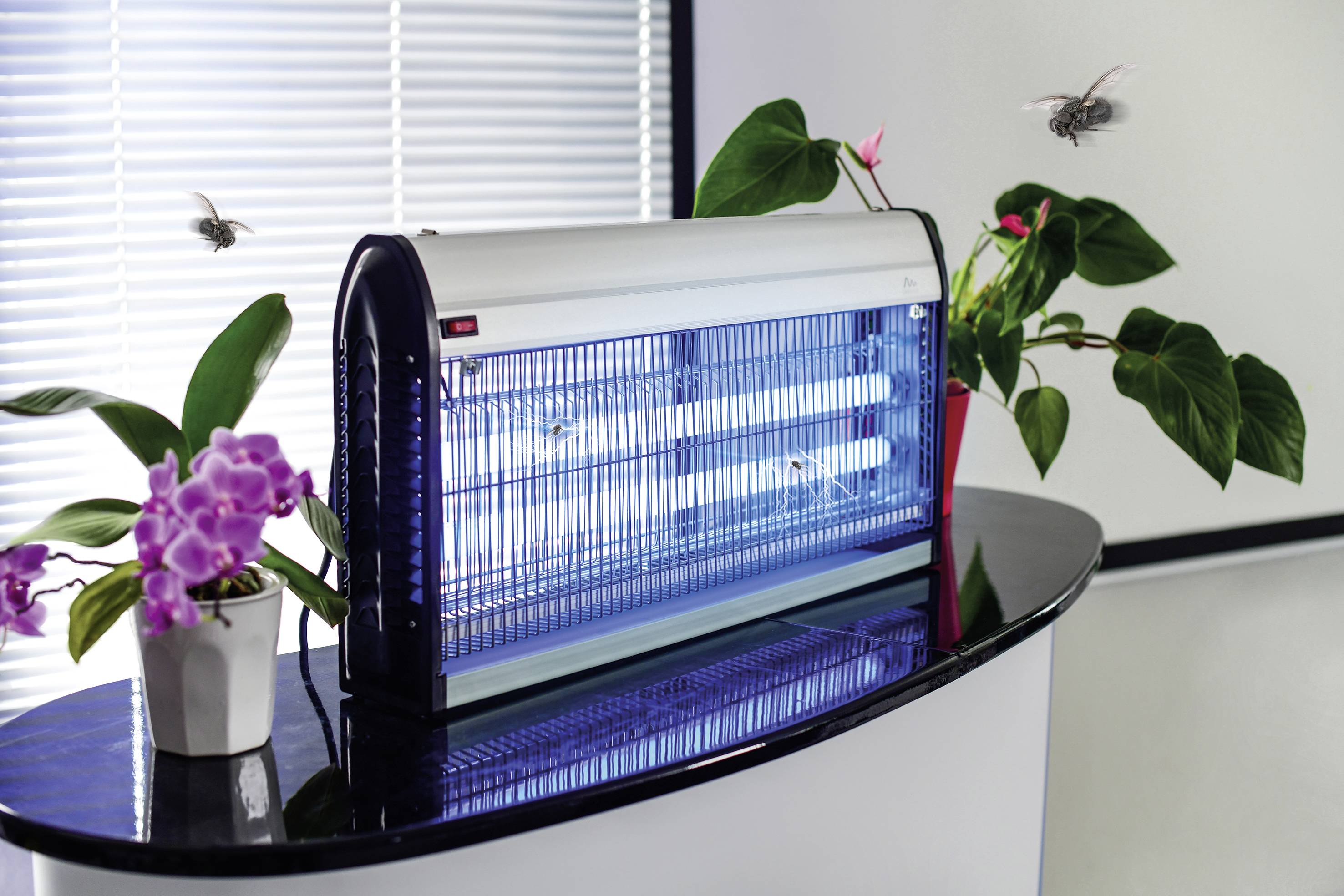 Elektrischer Insektenvernichter auf einem Tisch neben Pflanzen, umgeben von fliegenden Insekten, in einem hellen Raum.