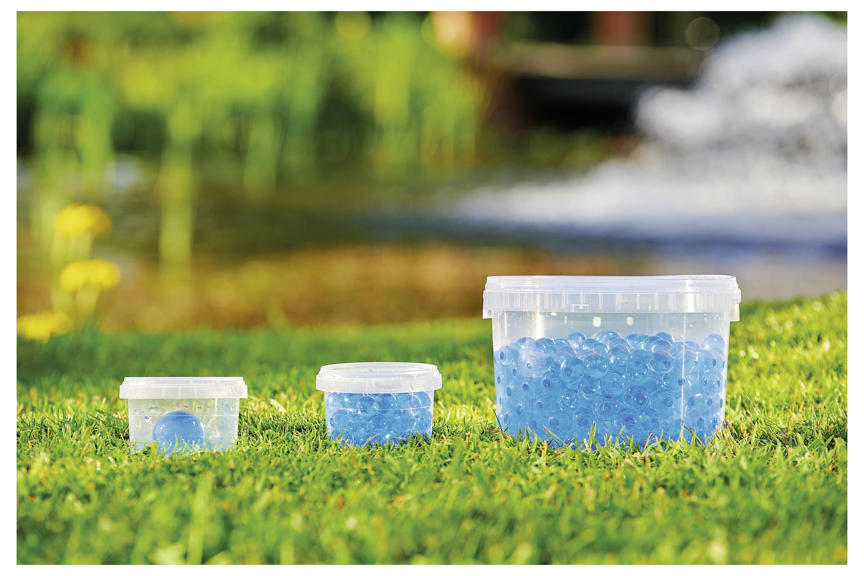 Drei Plastikbehälter unterschiedlicher Größe, gefüllt mit blauen Wasserbällen, stehen auf Gras, mit unscharfem natürlichen Hintergrund.