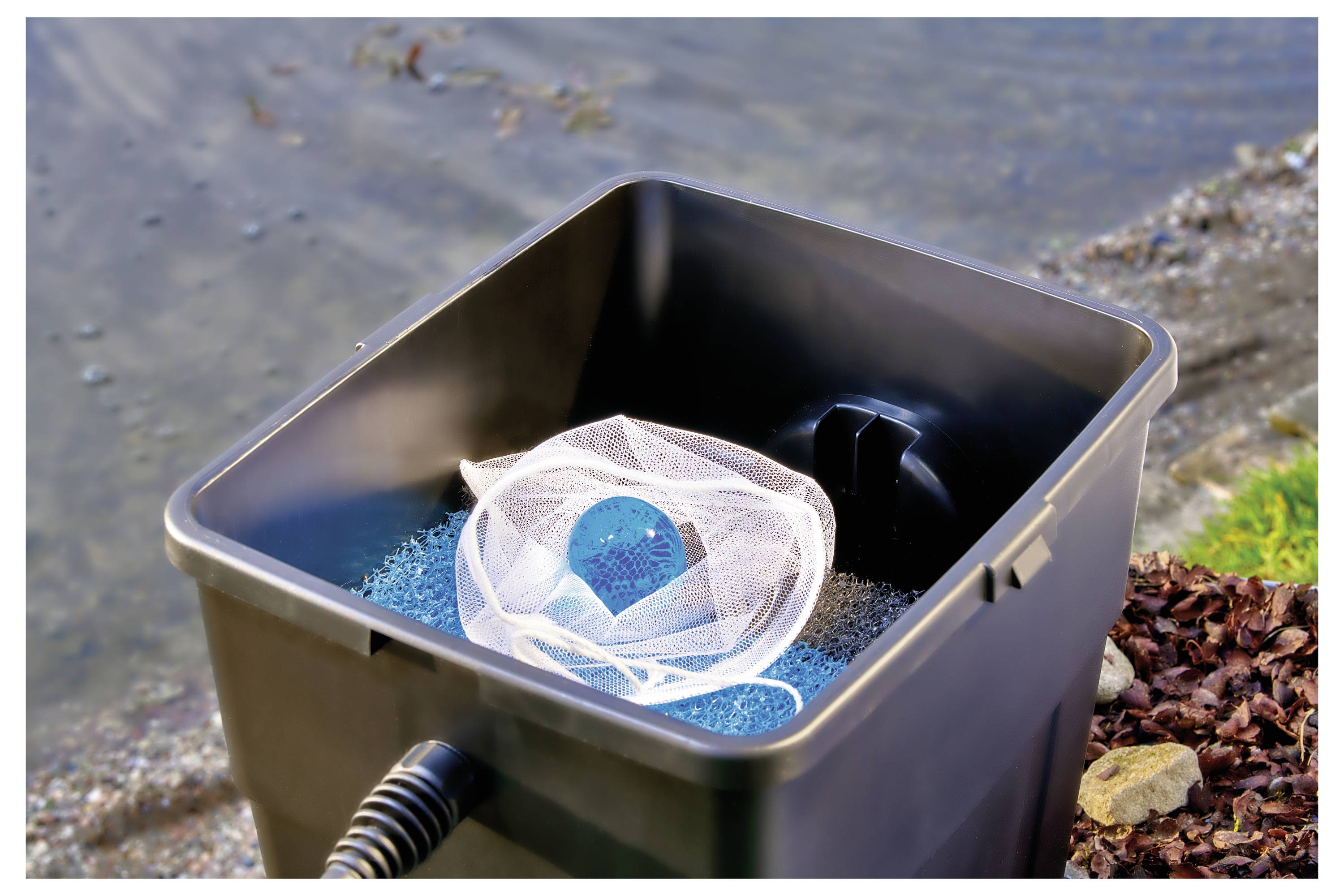 Eine Nahaufnahme eines Teichfilters an der Wasserkante, der einen Kescher und blaues Filtermaterial enthält. Steine und Wasser sind im Hintergrund zu sehen.