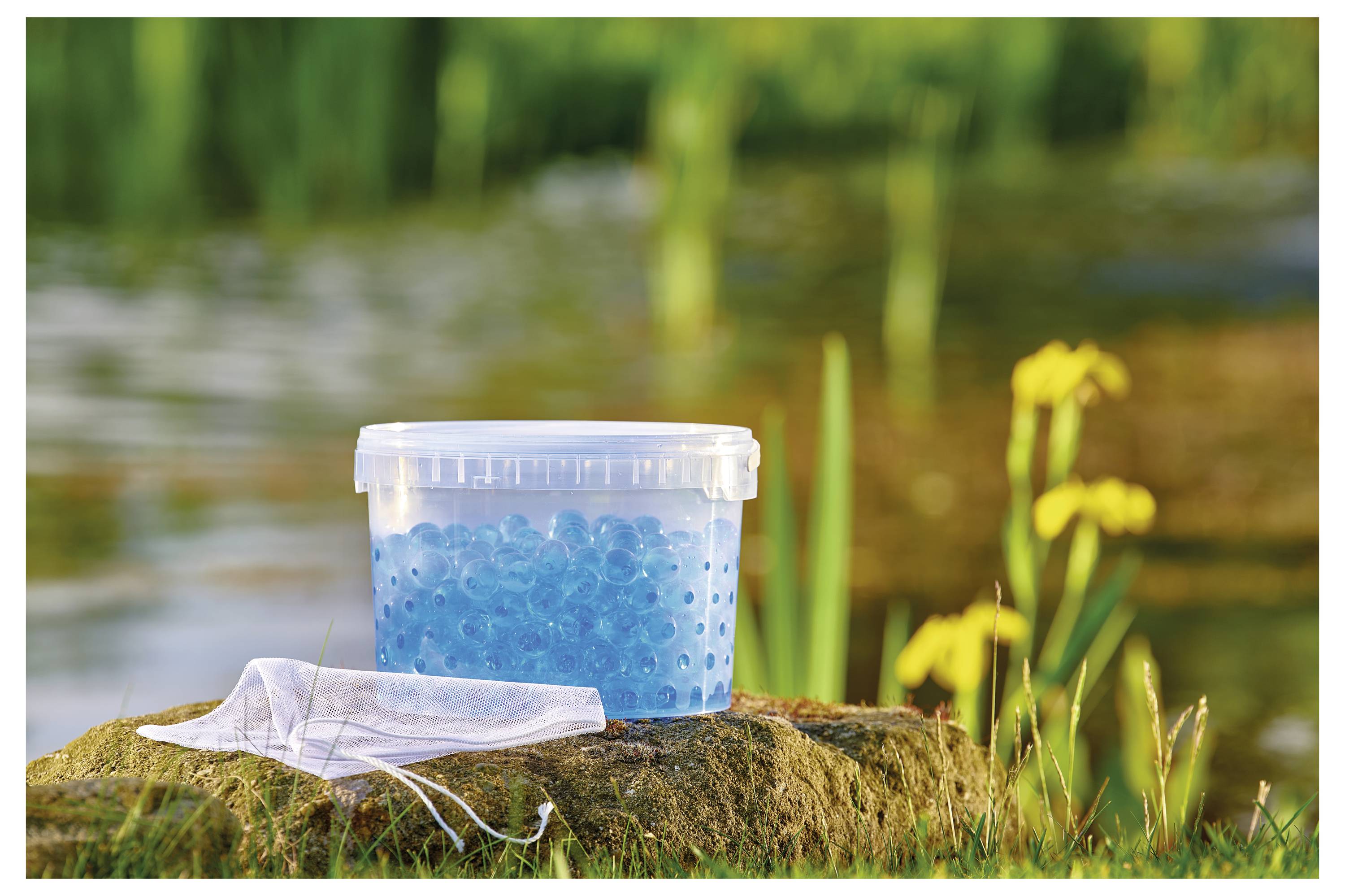 Ein Plastikbehälter mit blauen Wasserkugeln steht auf einem Stein neben einem Teich, umgeben von grünen Pflanzen und gelben Blumen. Eine Maske liegt daneben.