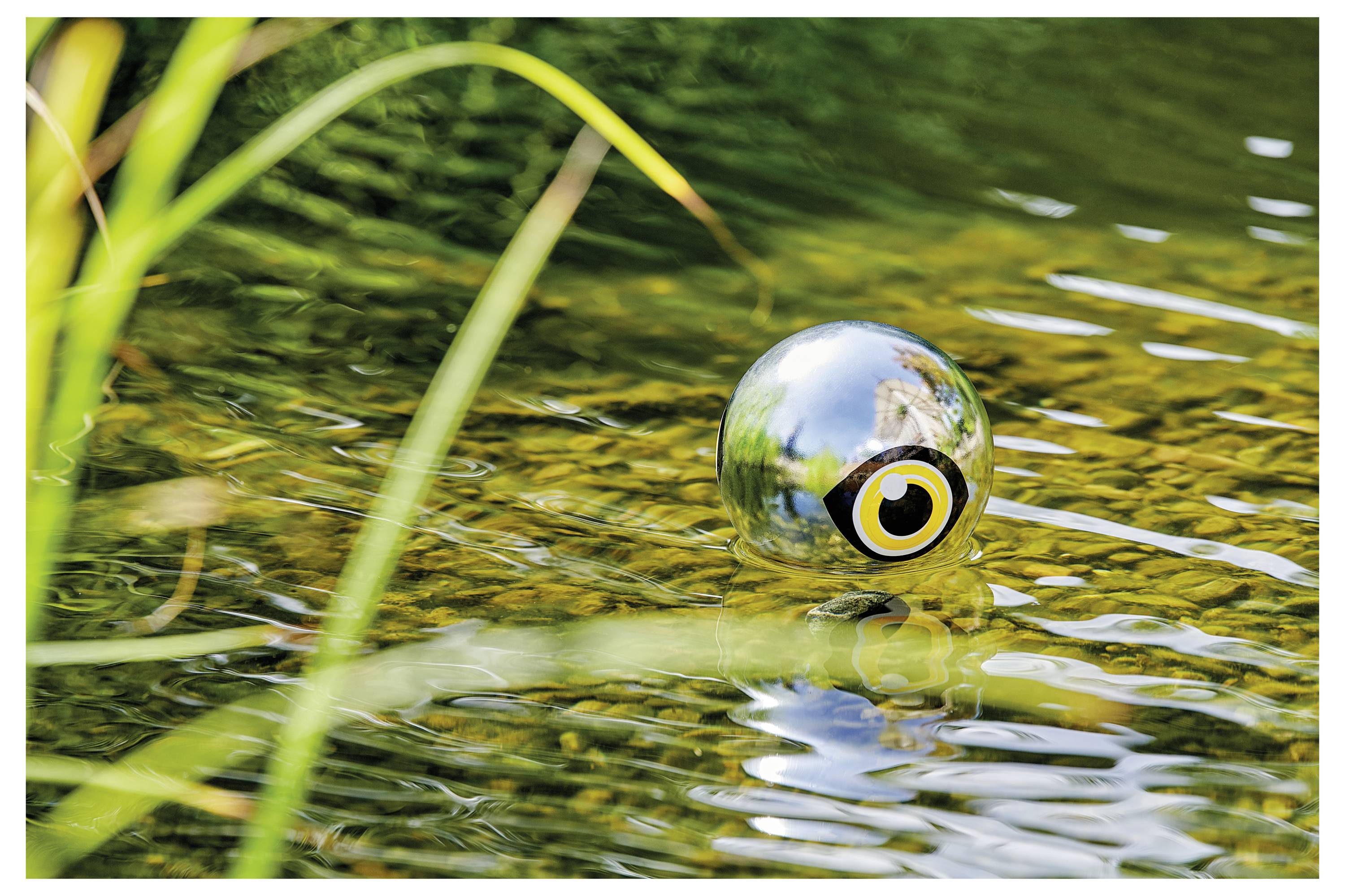 Eine reflektierende silberne Kugel mit Augendesign schwebt auf einem Teich, umgeben von Wasser und Grün, wodurch eine surreale visuelle Wirkung entsteht.