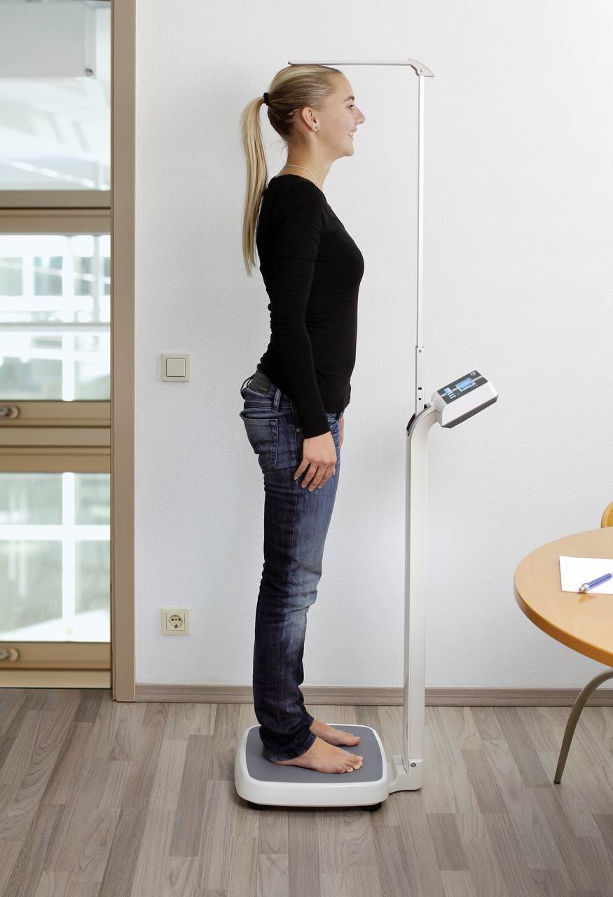 Eine Frau steht in einem Raum auf einer Waage mit Messstab, um ihre Körpergröße zu messen. Sie trägt ein schwarzes Oberteil und Jeans.