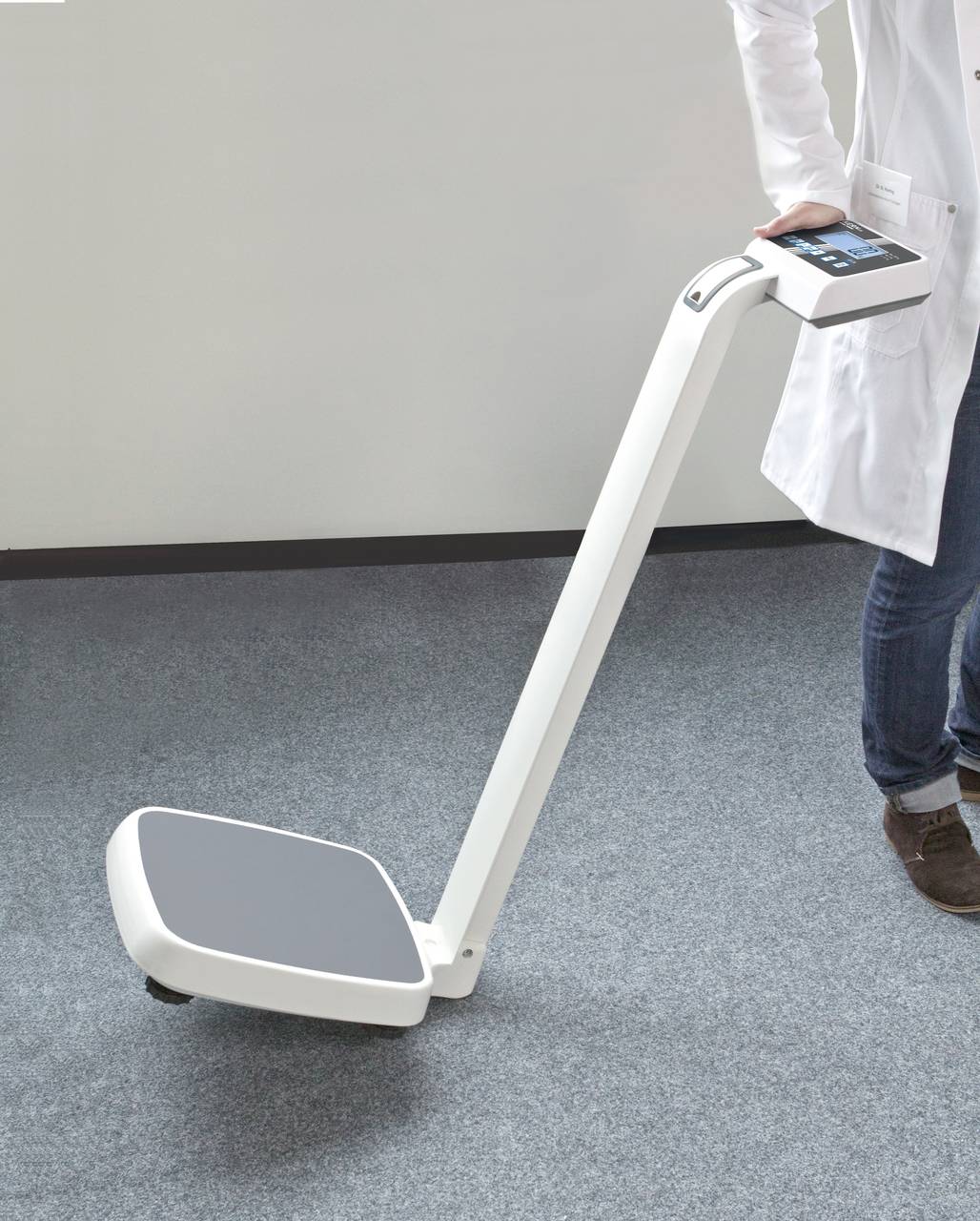 Eine Person in einem weißen Kittel hält eine moderne, tragbare Körperwaage mit einem Griff und einem digitalen Display auf Teppichboden.