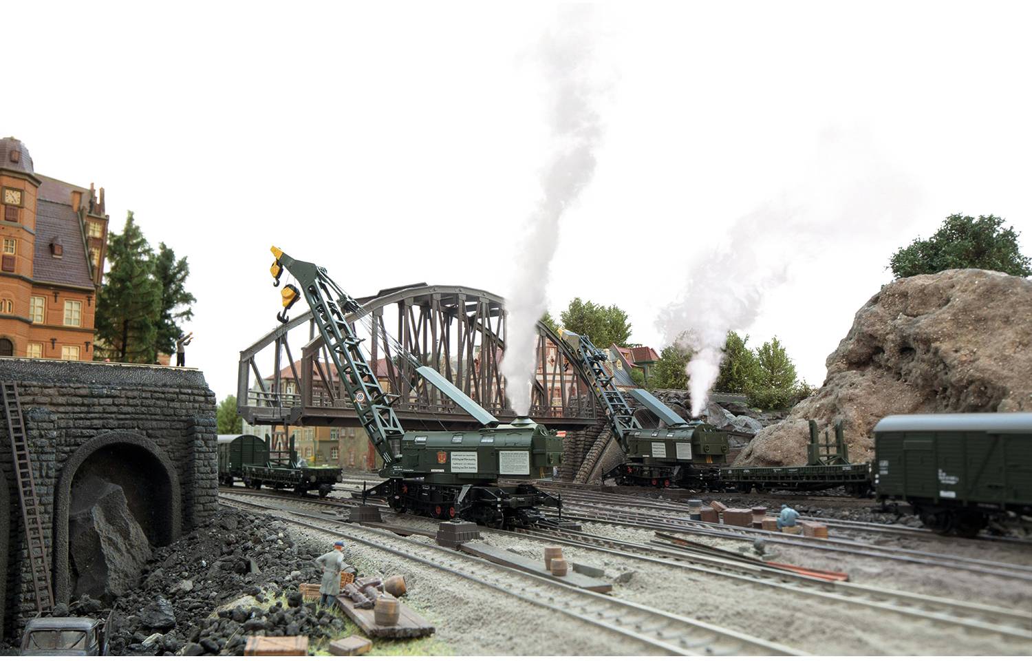 Modelleisenbahnszene mit zwei Dampflokomotiven, Kränen und einem Tunnel, vor einem Brückenhintergrund. Arbeiterfigur im Vordergrund.