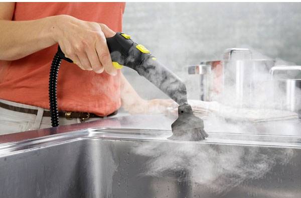 Eine Person reinigt eine Küchenspüle mit einem Dampfreiniger. Dampf steigt auf, während die Bürste über die Oberfläche geführt wird.