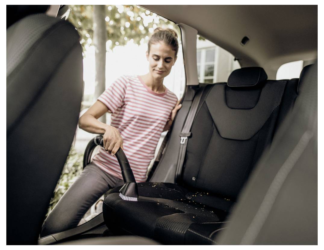 Eine Person in einem gestreiften Shirt saugt den Rücksitz eines Autos aus und entfernt Schmutz. Sonnenlicht fällt durch die Fenster und deutet auf eine Reinigungsaufgabe hin.