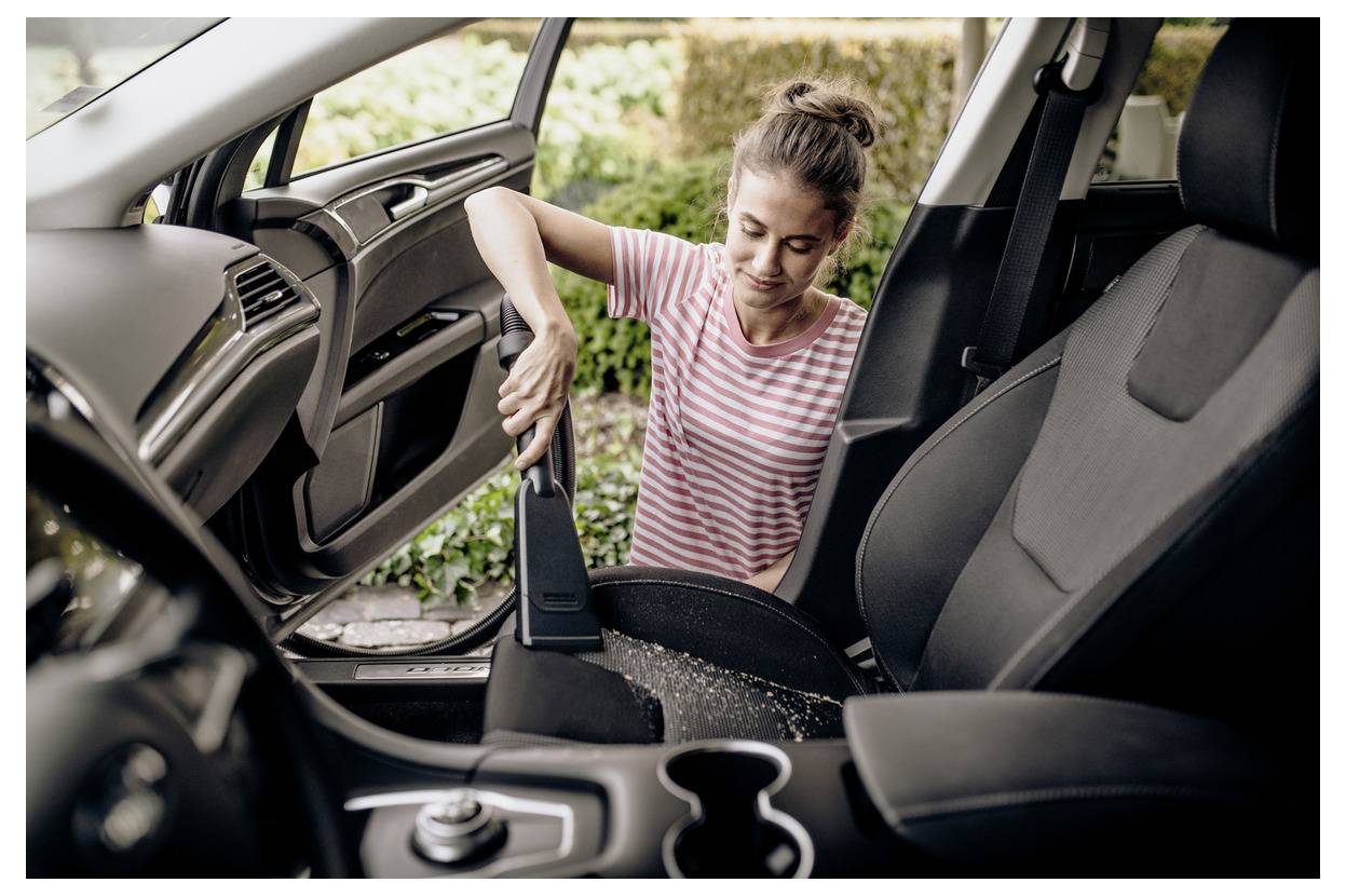 Eine Frau in einem gestreiften Shirt saugt den Beifahrersitz eines Autos aus und entfernt dabei Schmutz. Die Autotür ist geöffnet, und im Hintergrund ist Grünzeug zu sehen.