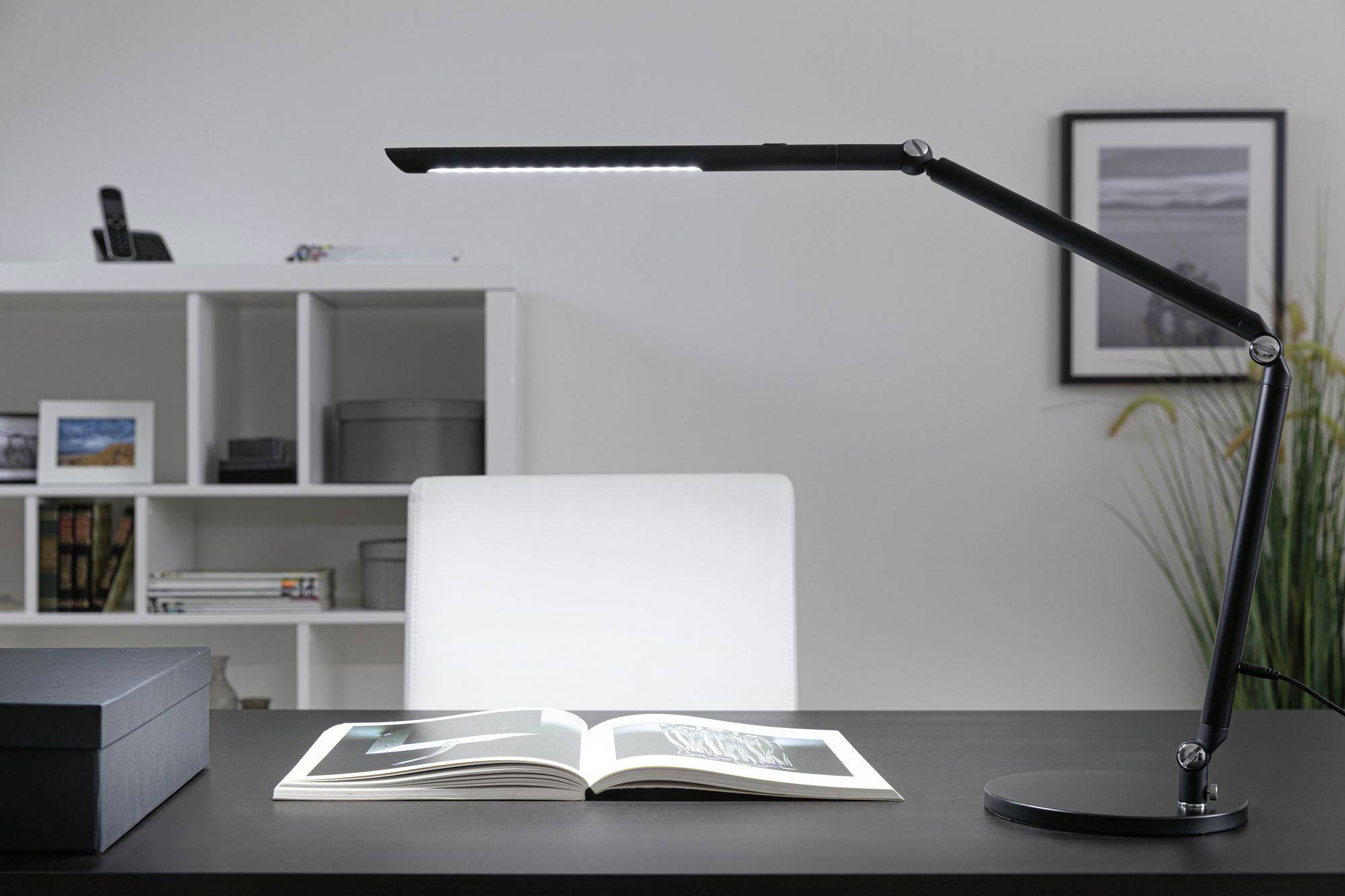 Schreibtisch mit weißem Stuhl, offenen Buch und schwarzer LED-Schreibtischlampe. Im Hintergrund ein Regal mit Büchern und Dekoration.