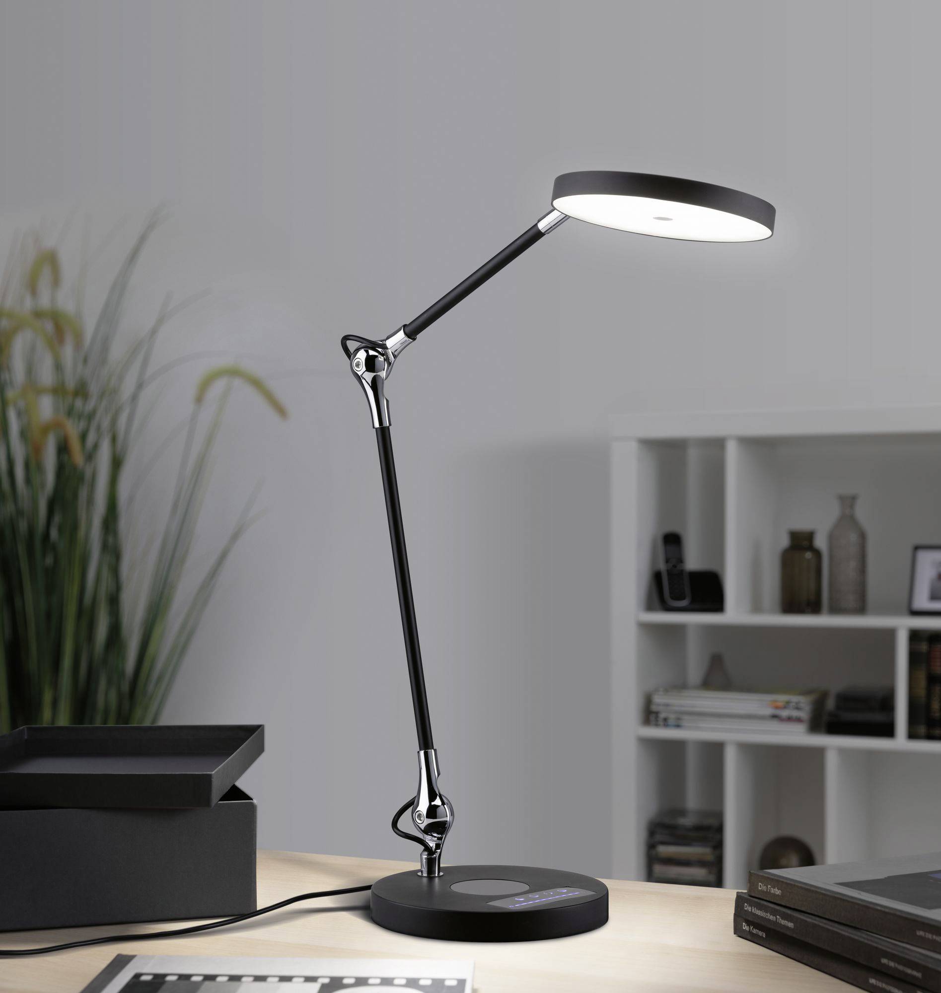 Eine moderne Schreibtischlampe leuchtet einen Arbeitsbereich aus. Im Hintergrund sind ein Bücherregal und eine Zimmerpflanze sichtbar.