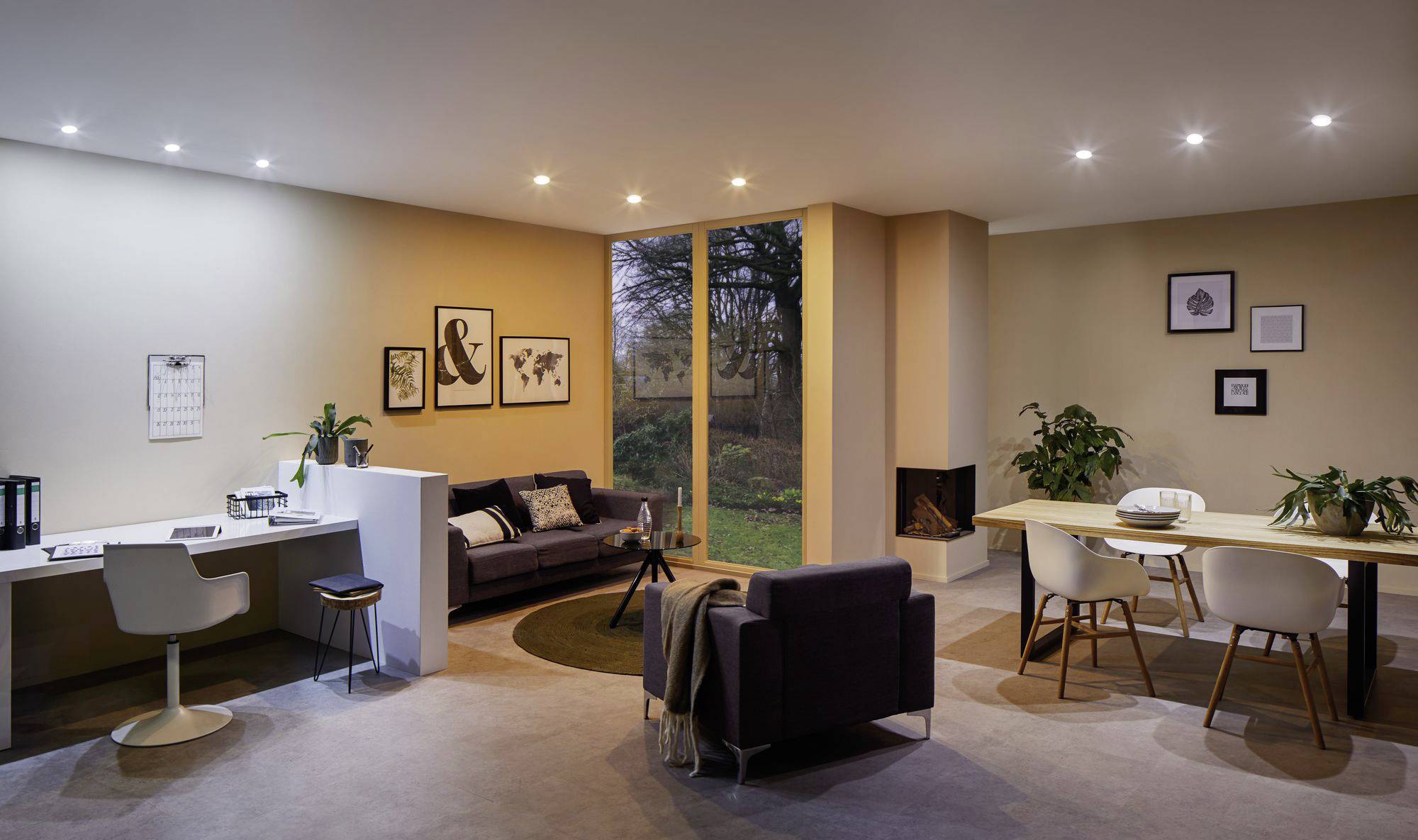 Ein modernes Wohnzimmer mit Schreibtisch, Sofa, Kamin und Esstisch. Große Fenster bieten Blick in den Garten. Skandinavisches Design.