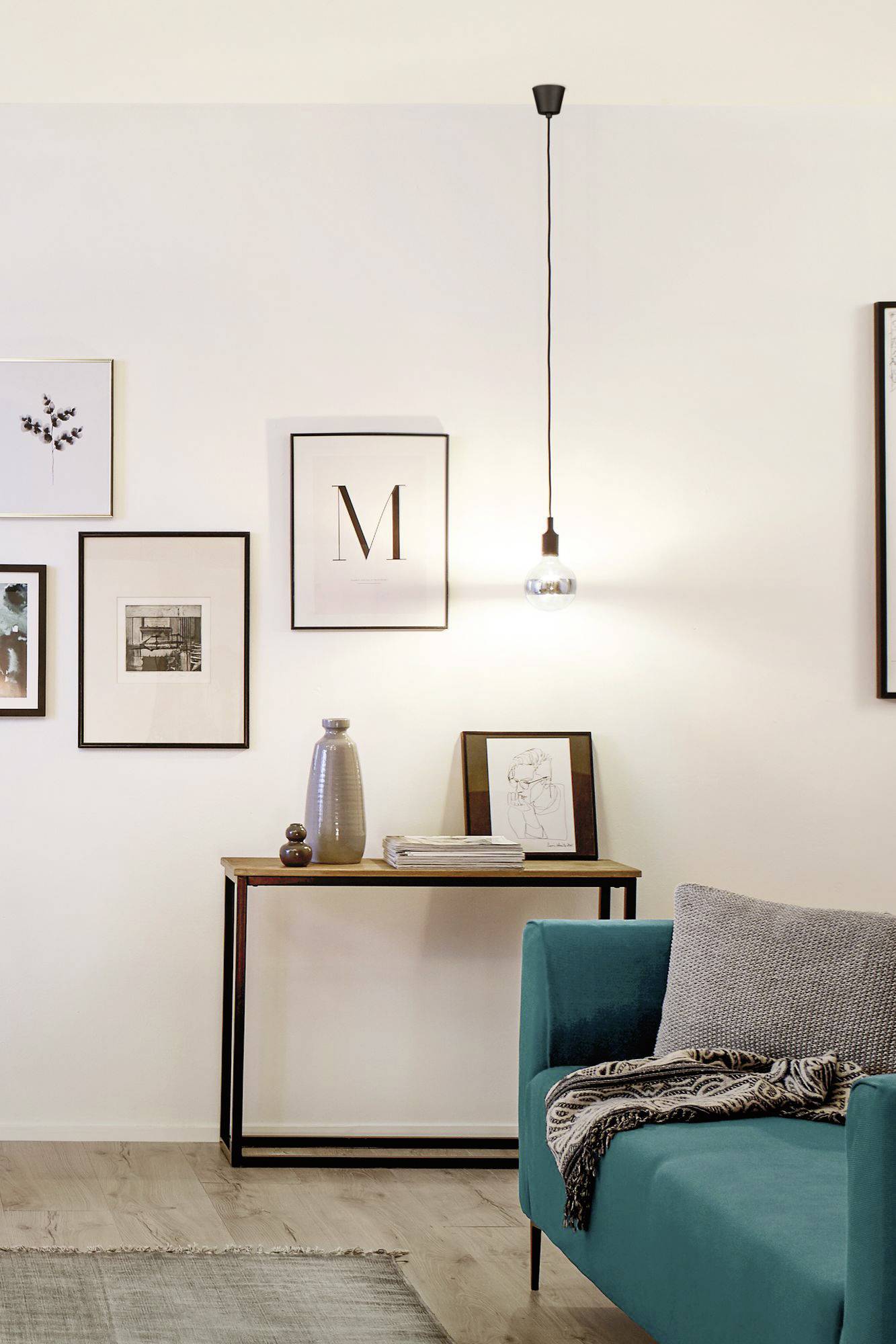 Ein modernes Wohnzimmer mit einem türkisfarbenen Sofa, auf dem eine graue Decke liegt. An der Wand hängen Bilderrahmen und eine Lampe leuchtet.