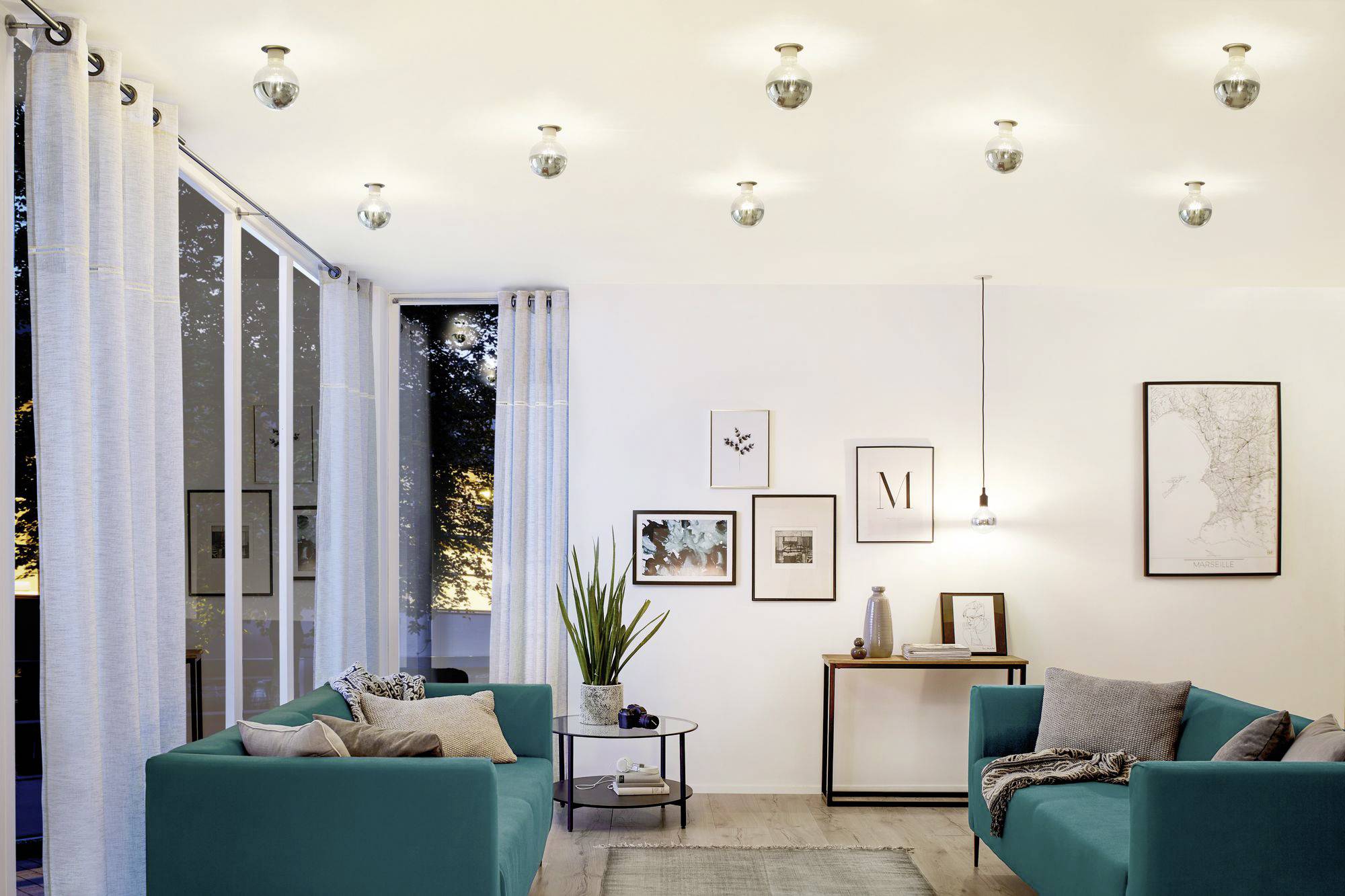 Wohnzimmer mit weißen Wänden, zwei türkisen Sofas, mehreren an der Decke angebrachten Lampen und dekorativen Bildern an der Wand.