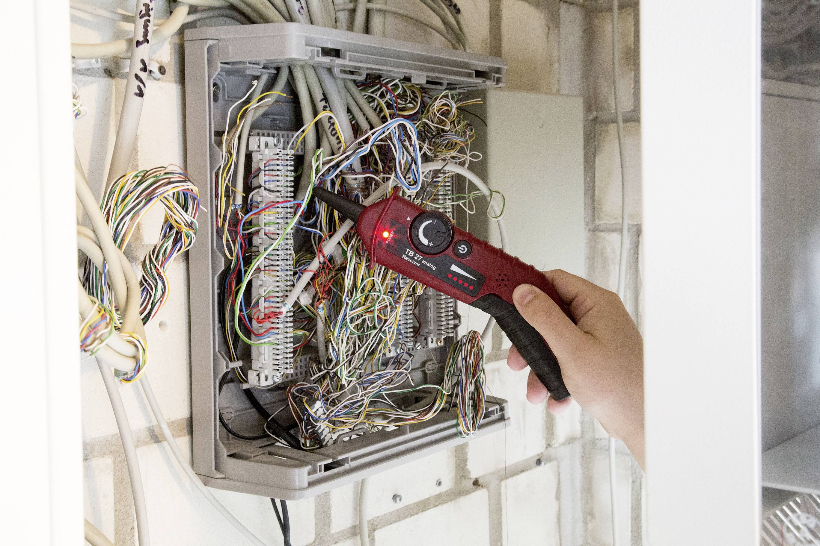 Eine Person testet mit einem Gerät Kabel in einer geöffneten Anschlussdose an einer Wand. Sichtbare Drahtverbindungen und Verbindungen.