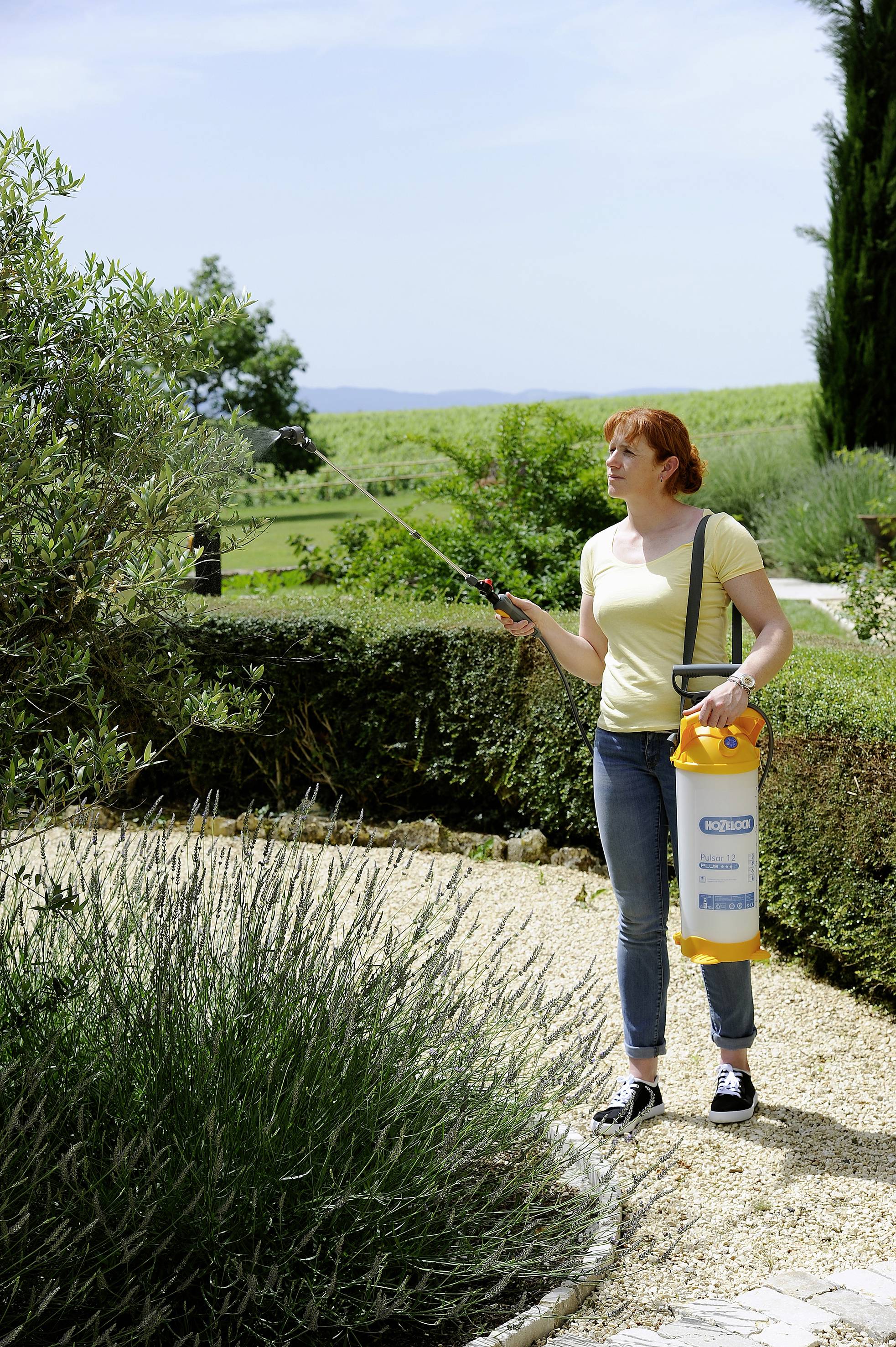 Frau mit gelbem T-Shirt sprüht Pflanzen in einem Garten mit einem Behälter für Pflanzenschutzmittel. Im Hintergrund weite Landschaft.
