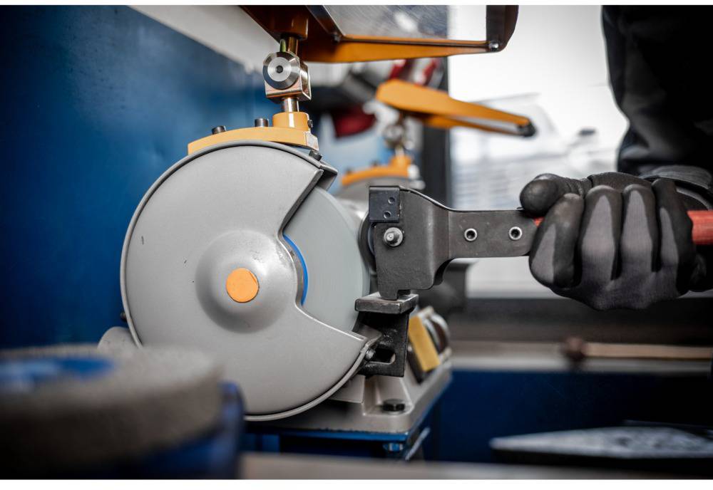 Ein Arbeiter schleift ein Werkzeug an einer Schleifmaschine. Er trägt dunkle Arbeitshandschuhe. Im Hintergrund sind Werkzeuge sichtbar.