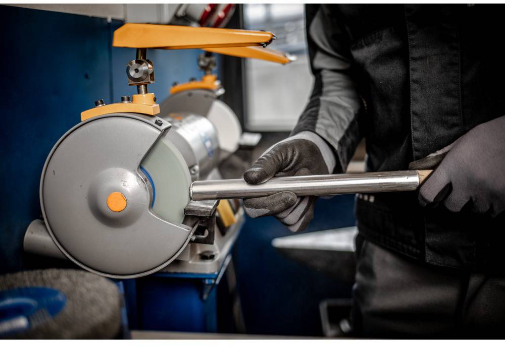 Eine Person schleift ein Metallwerkzeug mit einer Schleifmaschine in einer Werkstatt. Die Person trägt Sicherheitskleidung und Handschuhe.