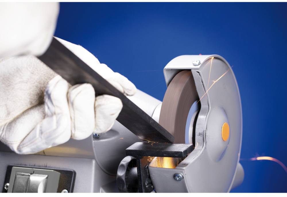 Person mit Handschuhen schleift Metallstab an einer Schleifmaschine, Funken sprühen.