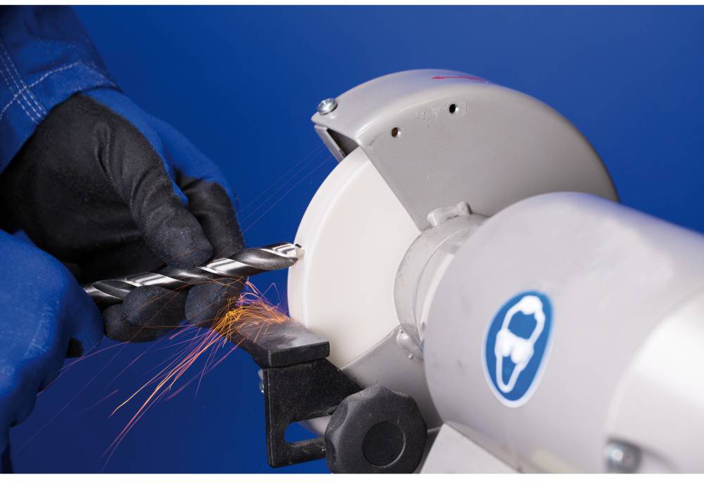 Person schleift einen Metallbohrer an einer Schleifmaschine. Funken fliegen beim Kontakt. Trägt Handschuhe für Sicherheit.