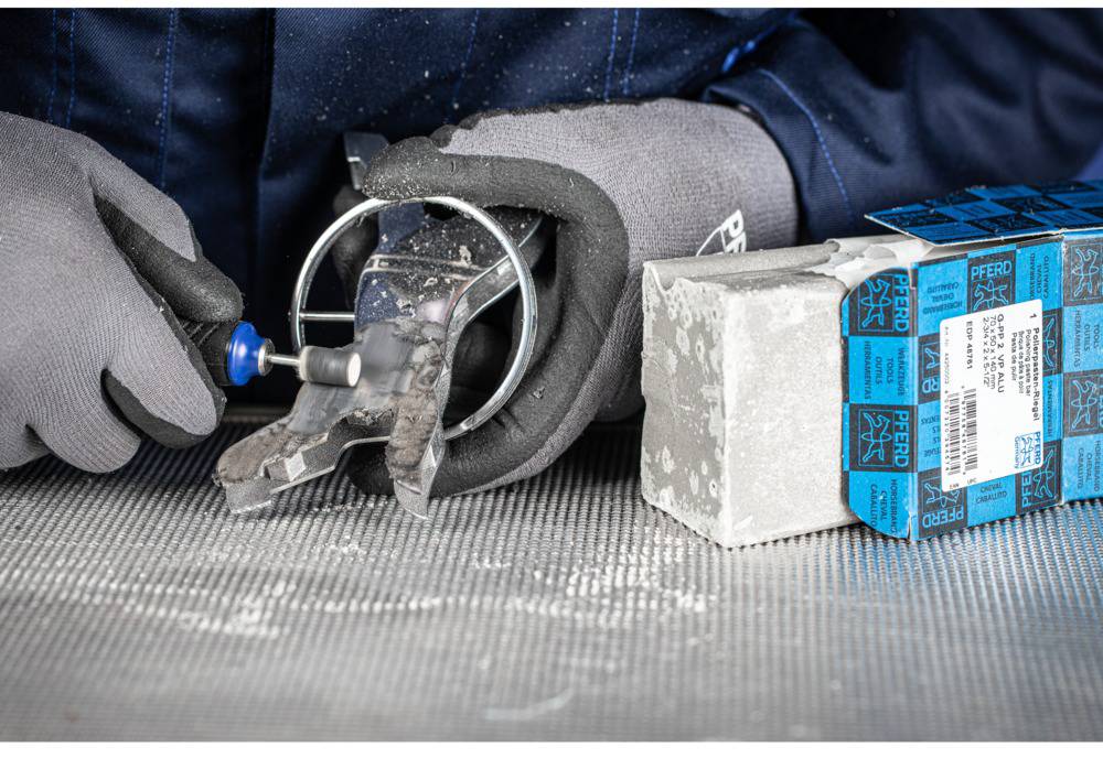 Nahaufnahme einer Person in Schutzhandschuhen, die mit einem rotierenden Werkzeug Metall bearbeitet. Rechts daneben ein Stapel mit Schachteln.