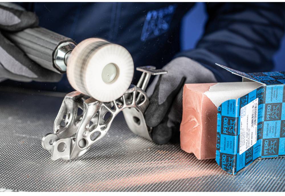 Eine Person poliert ein komplex geformtes Metallteil mit einer rotierenden Bürste. Daneben liegt eine geöffnete Verpackung mit Schleifmitteln.