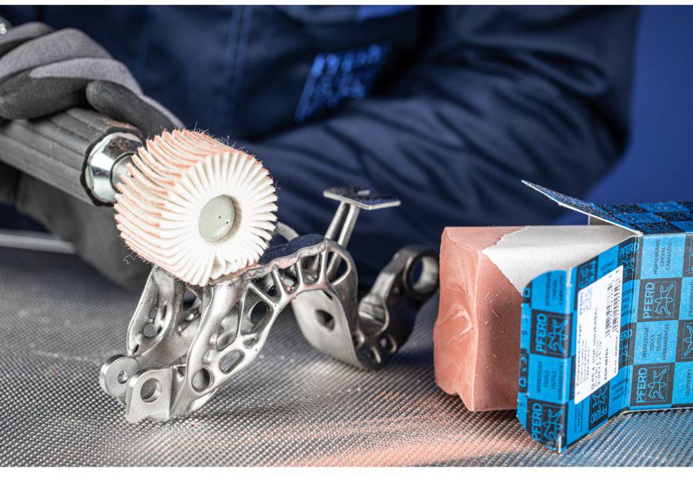 Eine Person poliert ein metallisches, komplexes Teil mit einer rotierenden Bürste. Daneben liegt ein blauer Schleifblock auf einem Tisch.