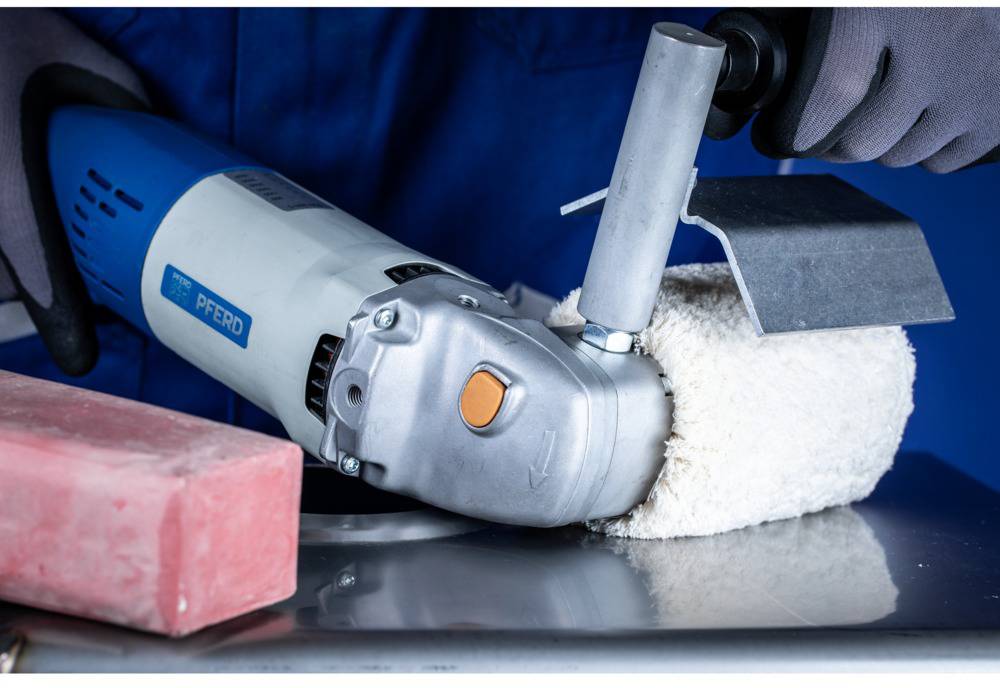Eine Person bearbeitet eine metallische Fläche mit einem Elektrowerkzeug und einem Polieraufsatz. Im Vordergrund sind ein roter Block und Schutzkleidung zu sehen.