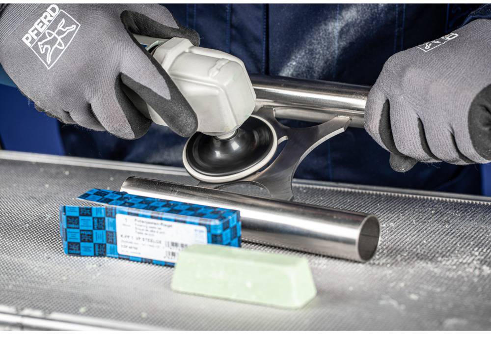 Eine Person poliert mit einer Maschine eine Metallstange. Im Vordergrund liegen eine Packung Schleifmittel und ein Schleifblock.