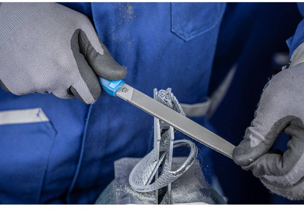 Eine Person in Arbeitskleidung bearbeitet mit einer Feile ein metallisches Werkstück. Nahaufnahme von Händen und Werkzeug.