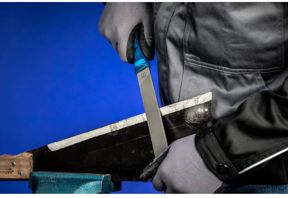 Eine Person in Arbeitskleidung und Handschuhen schärft mit einer Feile die Klinge eines Werkzeugs vor blauem Hintergrund.