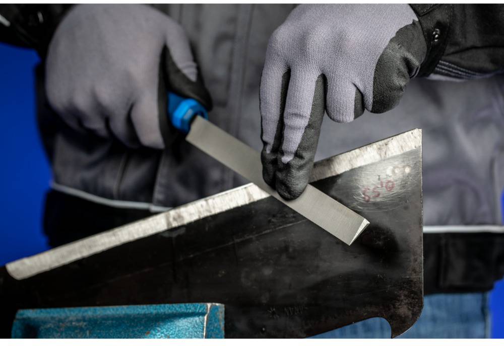 Person trägt graue Handschuhe und schleift eine Metallkante mit einer Feile. Der Hintergrund ist blau.