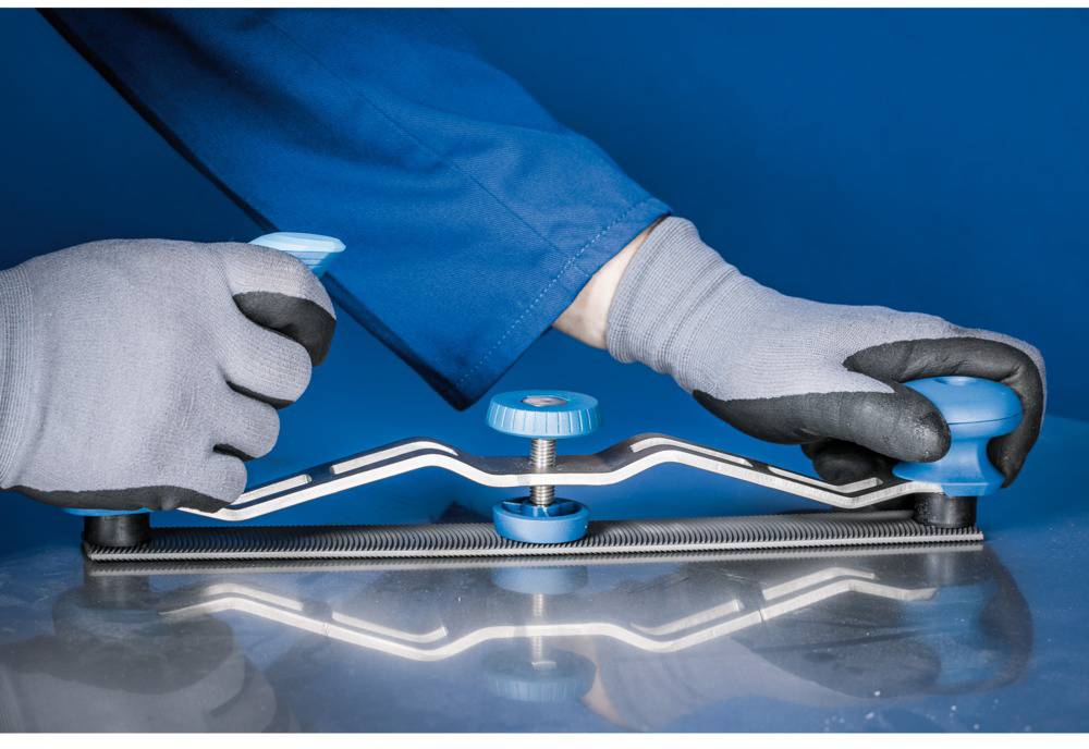 Eine Person in blauer Arbeitskleidung benutzt ein Handwerkzeug zur Metallbearbeitung. Das Werkzeug hat blaue Griffe und gleitet über eine Metalloberfläche.